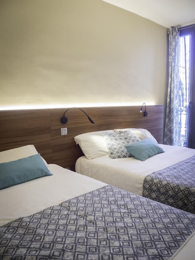 A cozy hotel room features two neatly arranged beds with patterned throws, wooden headboards, modern wall lamps, and soft natural light filtering through lace curtains, ideal for travelers seeking comfort and tranquility.