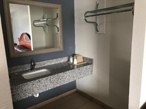 A cozy hotel bathroom features a sleek granite countertop sink, a stylish framed mirror reflecting a bed, and chrome towel racks, ideal for travelers seeking comfort and modern amenities.