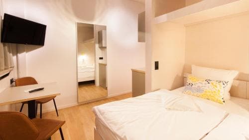 A cozy hotel room features a neatly made bed, a desk with chairs, and a mirror reflecting the space, creating a warm and inviting atmosphere ideal for travelers seeking comfort and functionality.