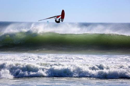 A windsurfer soars above crashing ocean waves under a clear blue sky, capturing the thrill of coastal adventure sports in a vibrant seascape ideal for adrenaline-seeking travelers.