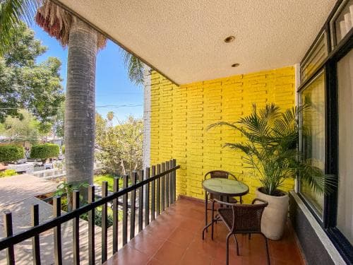 A cozy balcony with a vibrant yellow brick wall, tiled floor, and lush greenery offers a serene retreat, perfect for travelers seeking relaxation in a sunny urban setting.