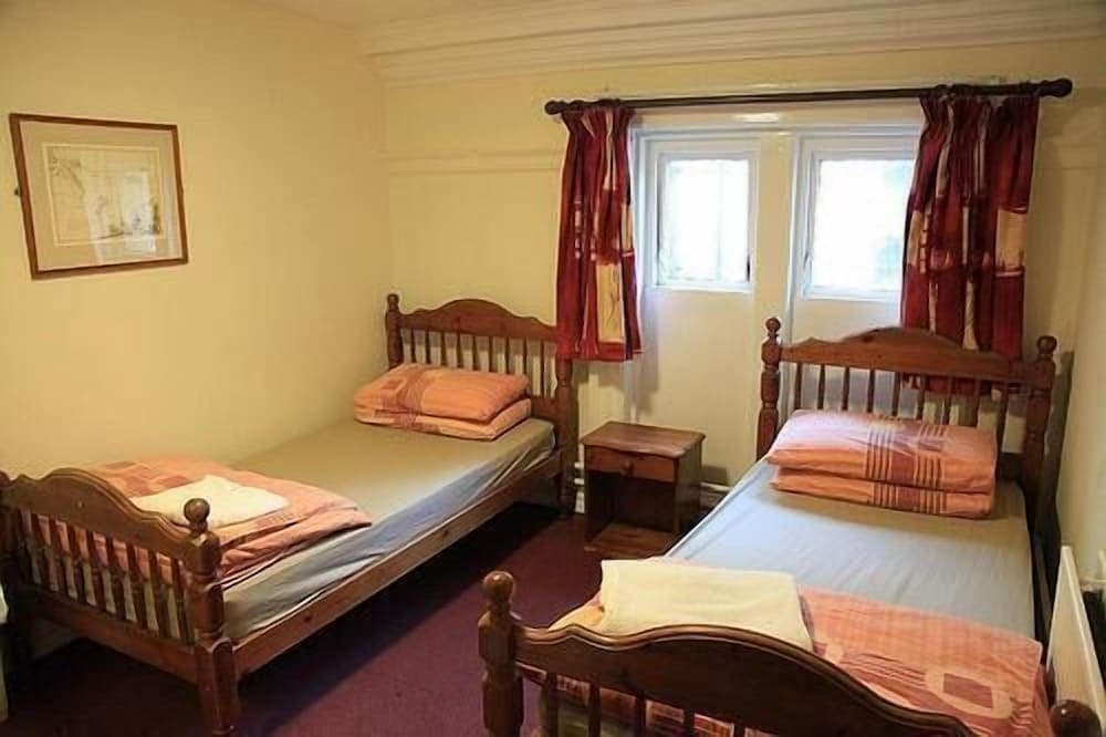 A cozy guesthouse bedroom features three wooden single beds with neatly folded blankets, red curtains framing a bright window, and warm yellow walls, ideal for budget travelers seeking comfort.