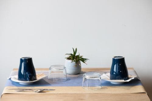 A serene minimalist dining setup features two blue cups, clear glasses, a small potted plant, and a striped tablecloth on a wooden table, ideal for travelers seeking a peaceful café or home dining experience.