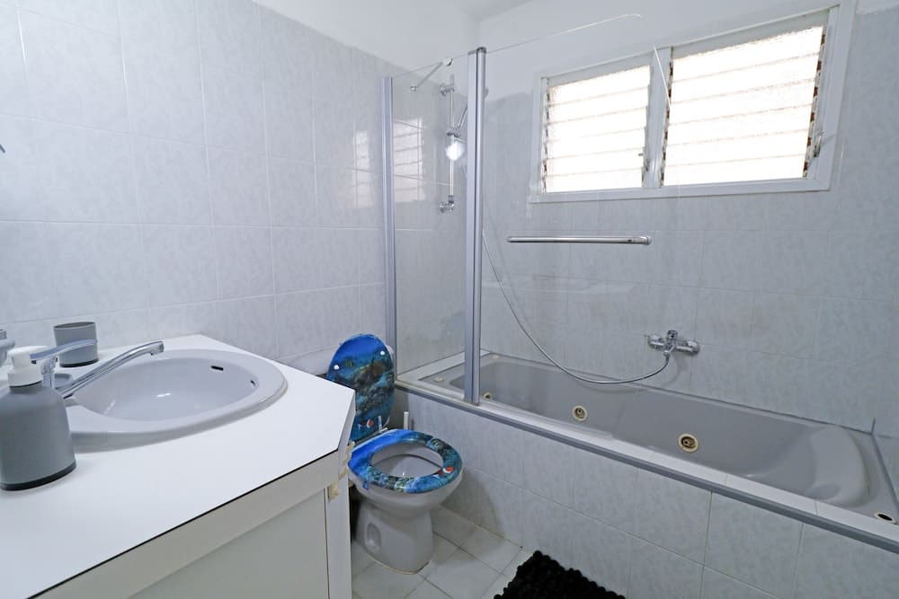 A clean, modern bathroom with white tiled walls, a bathtub with a glass shower screen, a sink with toiletries, and a toilet with a colorful seat cover, ideal for travelers seeking comfortable accommodations.