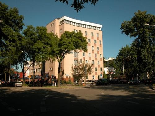 A serene urban street scene featuring a multi-story residential building surrounded by lush green trees under a clear blue sky, ideal for travelers seeking a peaceful city stay with easy access to nature.