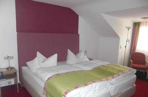 A cozy hotel room features a neatly made bed with white linens and a green throw blanket, set against a vibrant magenta headboard, complemented by soft lighting and a comfortable chair, ideal for travelers seeking a relaxing stay.