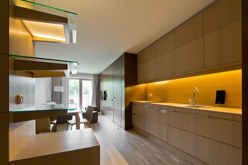 A modern, minimalist hotel suite features sleek wooden cabinetry, warm ambient lighting, and a cozy dining area overlooking a serene outdoor view, ideal for travelers seeking comfort and style.