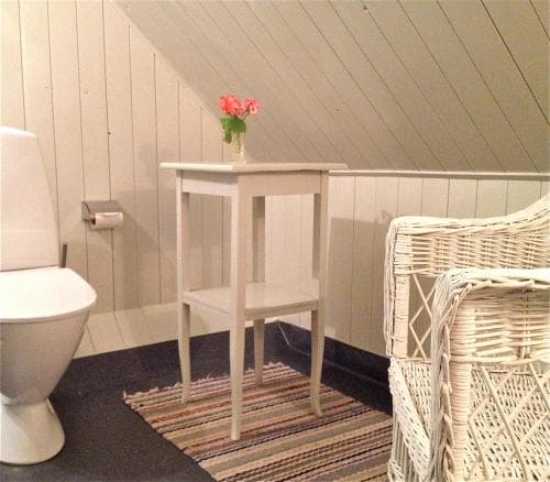 A cozy attic bathroom with white wooden walls, a small table holding pink flowers, a wicker chair, and a striped rug creates a serene and inviting space perfect for a relaxing getaway.