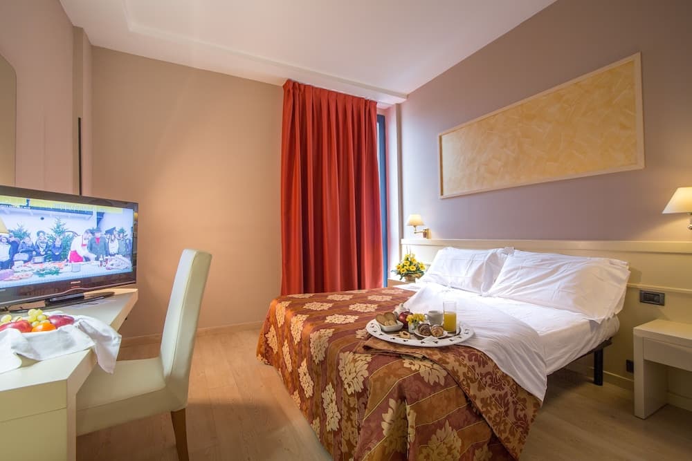 A cozy hotel room features a neatly made bed with a floral-patterned blanket, a vibrant red curtain, a desk with a TV displaying a festive scene, and a breakfast tray, ideal for travelers seeking comfort and convenience.
