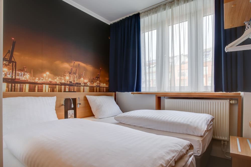 A cozy hotel room features twin beds with crisp white linens, a vibrant harbor mural on the wall, large windows with blue curtains, and a minimalist design, ideal for travelers seeking comfort and a touch of local scenery.