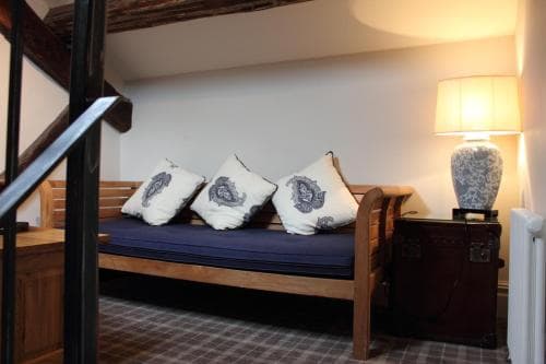 A cozy nook features a wooden bench adorned with patterned cushions under a staircase, illuminated by a vintage lamp on a side table, ideal for travelers seeking a quaint and serene accommodation spot.