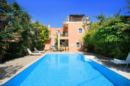 A serene Mediterranean villa courtyard features a vibrant blue pool surrounded by lush greenery, lounge chairs, and a charming pink building, ideal for a relaxing vacation retreat.