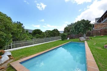 A serene backyard pool surrounded by lush greenery and a white picket fence under a bright blue sky, ideal for relaxing summer getaways.