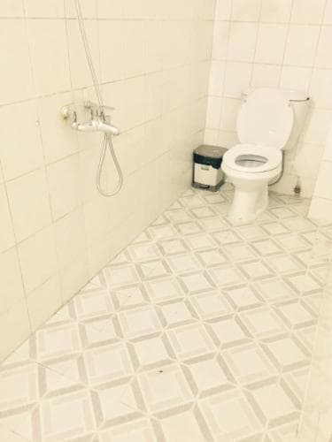 A clean, minimalistic bathroom with white tiled walls and floor features a toilet, handheld shower, and small trash bin, ideal for travelers seeking basic hygiene facilities in compact spaces.
