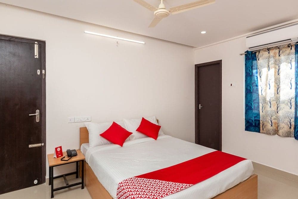 A cozy hotel room features a neatly made bed with red accents, tree-patterned curtains, and modern amenities, ideal for travelers seeking comfort and convenience.