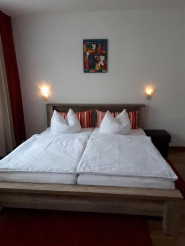 A cozy hotel room features a neatly made double bed adorned with white linens and red pillows, set against a minimalist backdrop with warm wall sconces and a vibrant abstract painting, ideal for travelers seeking comfort and relaxation.