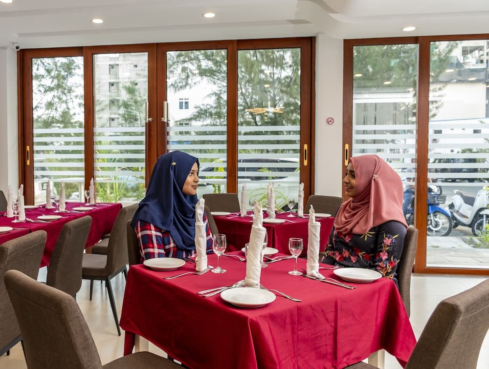 Two individuals in hijabs share a warm conversation at a cozy restaurant with red tablecloths, large windows offering a serene outdoor view, ideal for travelers seeking cultural dining experiences.