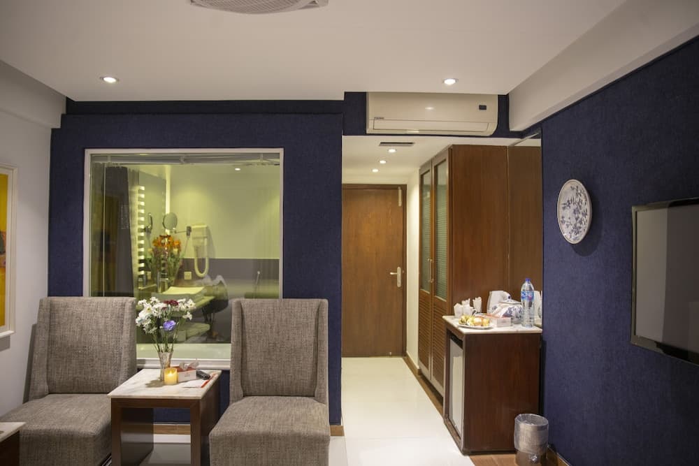 A cozy hotel room features a modern seating area with gray chairs, a small table adorned with flowers, a bathroom visible through a glass partition, and warm lighting that creates an inviting atmosphere ideal for travelers seeking comfort and relaxation.