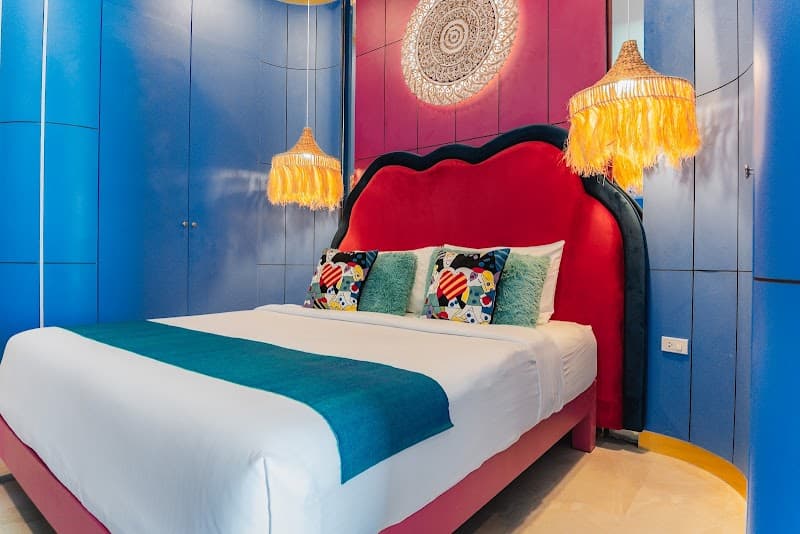 A vibrant bedroom at LOVE Beach Club Koh Samui features bold blue walls, a striking red bed adorned with colorful pillows, and unique tassel pendant lights, creating an inviting and stylish retreat for travelers seeking a luxurious stay.