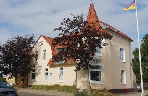 A charming two-story house with a red-tiled roof and beige walls stands amidst greenery under a partly cloudy sky, featuring a German flag and ideal for a quaint European getaway.