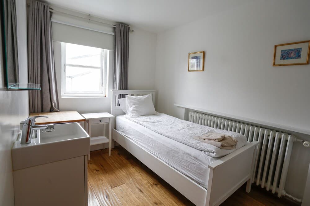 A cozy single bedroom with white furnishings, wooden flooring, a small sink, and framed artwork exudes a serene atmosphere, ideal for travelers seeking a minimalist and tranquil accommodation experience.
