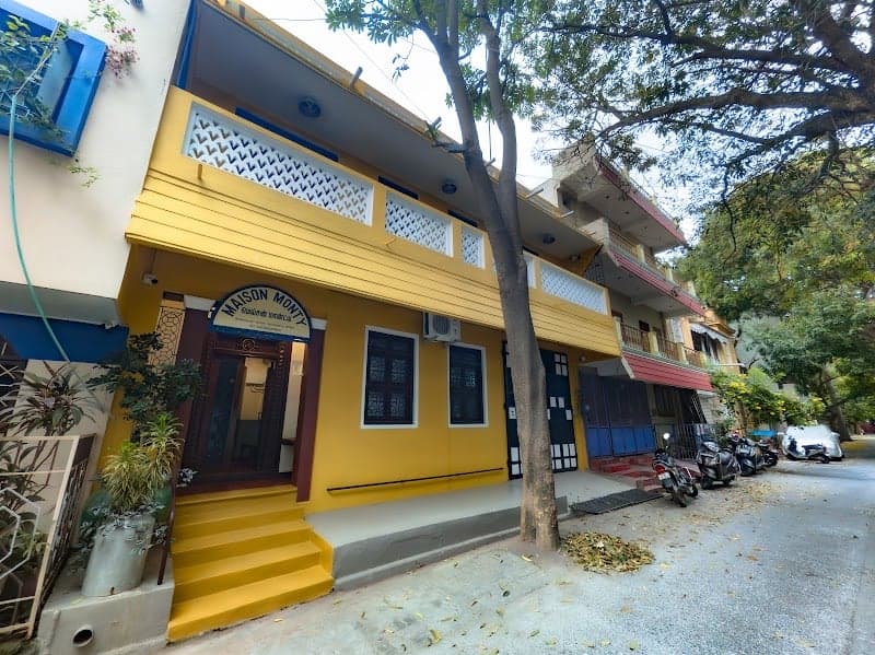 A vibrant yellow two-story building named "Maison Monty" stands invitingly on a quiet street in Puducherry, India, surrounded by lush greenery and motorbikes, ideal for travelers seeking a cozy and colorful accommodation experience.