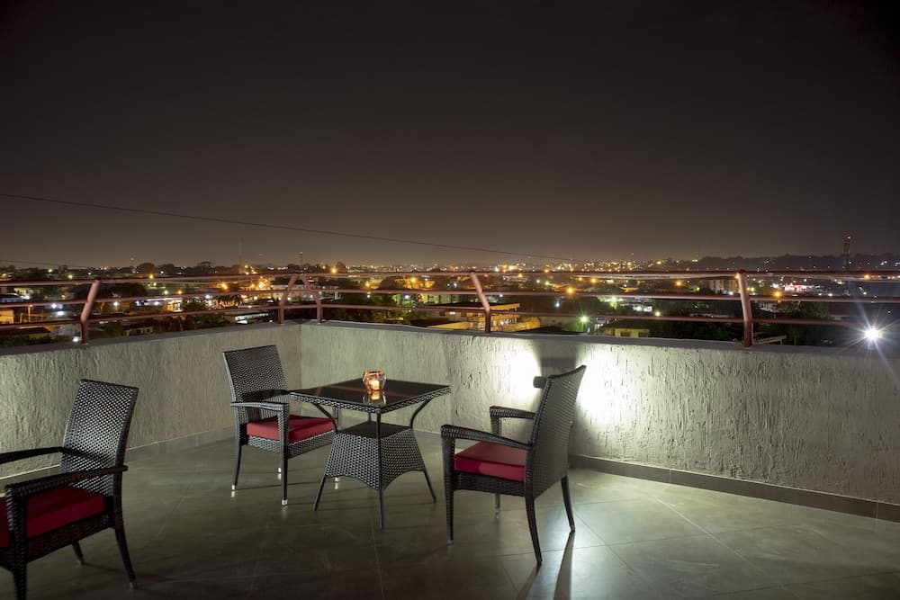 A cozy rooftop terrace at night features wicker furniture with red cushions, a candle-lit table, and a serene view of twinkling city lights, ideal for romantic getaways or evening relaxation.