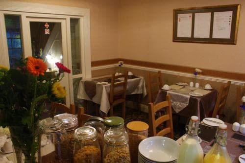 A cozy café interior features wooden chairs, tables set with white linens, vibrant flower arrangements, and jars of cereals on display, creating a warm and inviting atmosphere perfect for a leisurely breakfast or brunch outing.