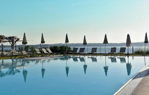 A serene poolside scene with lounge chairs and closed umbrellas reflecting in the calm water, set against a tranquil lakeside backdrop, ideal for a relaxing vacation getaway.