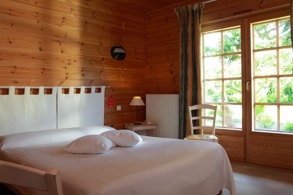 A cozy cabin bedroom with wooden walls, a neatly made bed, a chair by the window, and lush greenery outside, ideal for a serene getaway retreat.