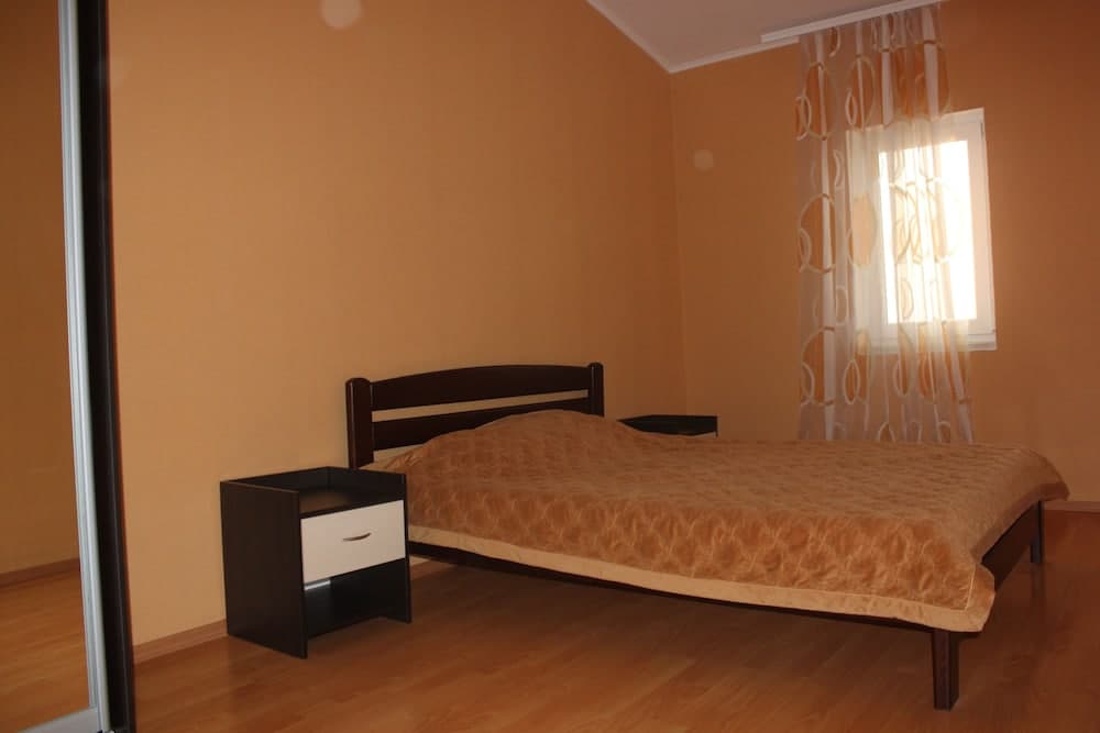 A cozy bedroom with warm orange walls, wooden flooring, a neatly made bed with an orange quilt, patterned curtains letting in soft light, and simple furniture, ideal for travelers seeking a comfortable and budget-friendly accommodation.