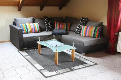 A cozy attic living space features a modern glass coffee table on a geometric rug, surrounded by comfortable sofas adorned with colorful pillows, ideal for a relaxing retreat or vacation rental.