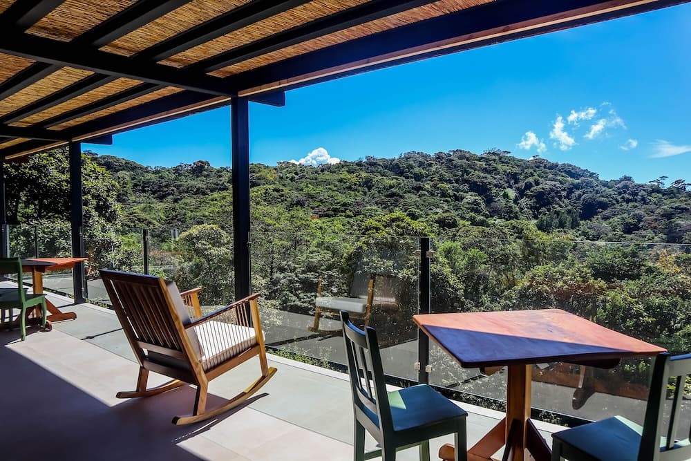 A serene outdoor patio with wooden rocking chairs and tables overlooks lush green hills under a clear blue sky, ideal for travelers seeking relaxation in a tranquil natural setting.