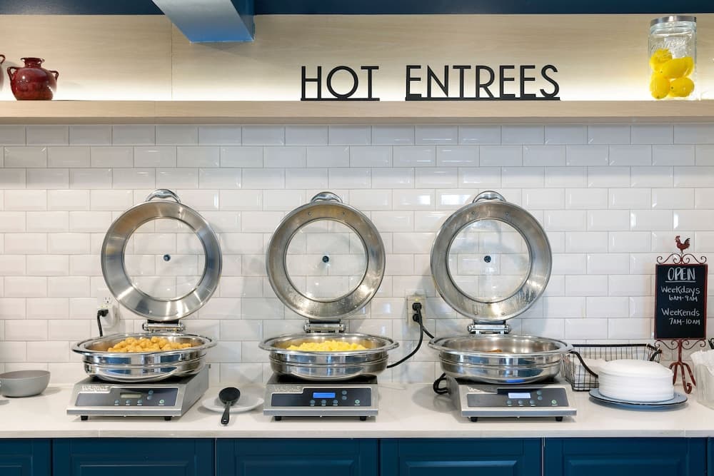 A cozy café's hot entrees station features stainless steel warming trays on digital scales against a white tiled wall, inviting travelers to enjoy freshly prepared meals in a welcoming atmosphere.
