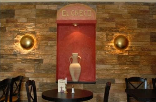 A cozy café corner features a rustic stone wall adorned with golden sconces and a red alcove displaying an elegant vase under the sign "EL GRECO," ideal for travelers seeking a charming spot to unwind.