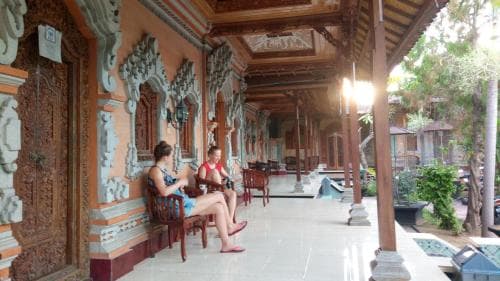 Visitors relax on ornate wooden chairs under the intricate carvings of a traditional Balinese temple veranda, capturing a serene cultural experience ideal for heritage tourism.