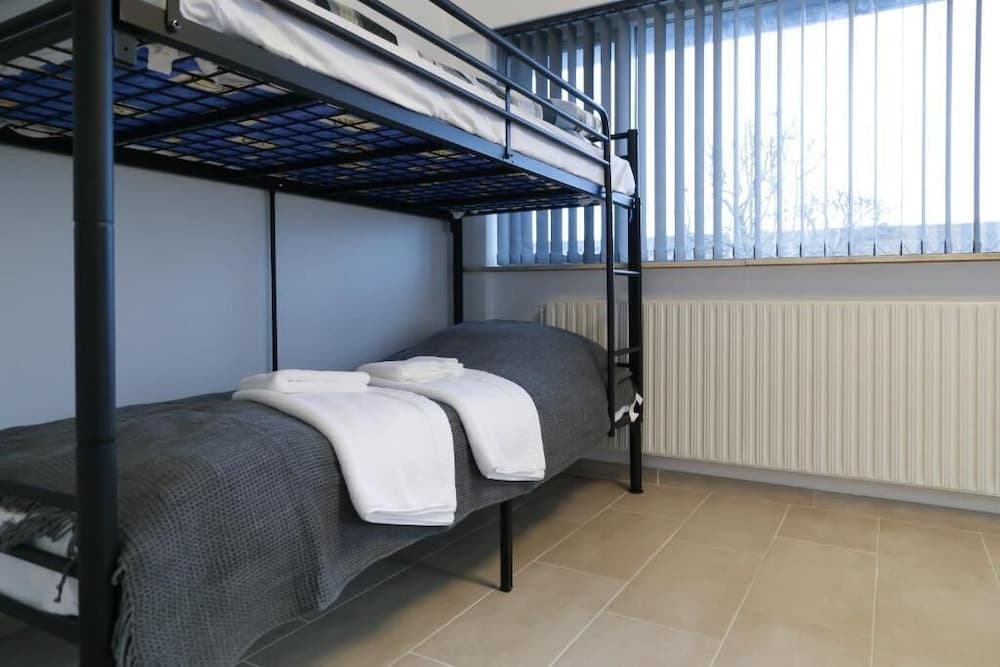 A cozy hostel room features a neatly arranged bunk bed with gray blankets and white towels, set against a window with vertical blinds, ideal for budget travelers seeking simple accommodations.