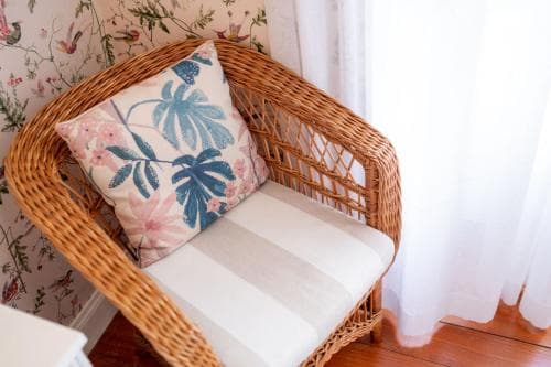 A cozy wicker chair adorned with a floral cushion sits invitingly in a charming room with botanical wallpaper, perfect for a relaxing getaway or home interior inspiration.