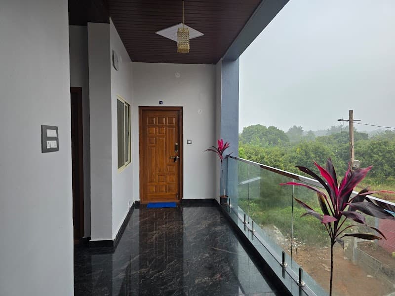 A serene balcony at Lakshmi Villa Hampi offers a tranquil view of lush greenery, featuring polished floors, glass railings, vibrant plants, and a cozy indoor-outdoor ambiance ideal for travelers seeking relaxation in Karnataka.