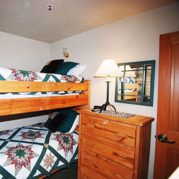 A cozy bedroom with wooden bunk beds adorned in quilted star-patterned bedding, a warm-toned dresser topped with a lamp, and a mirror reflecting the serene ambiance, ideal for family-friendly vacation rentals.