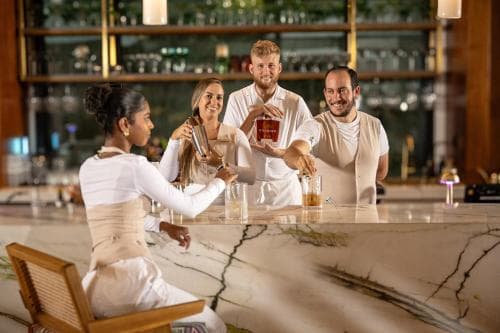 In a cozy bar setting with warm lighting and stocked shelves, four smiling individuals gather around a marble counter, enjoying drinks and conversation, ideal for capturing the essence of social gatherings and nightlife experiences.