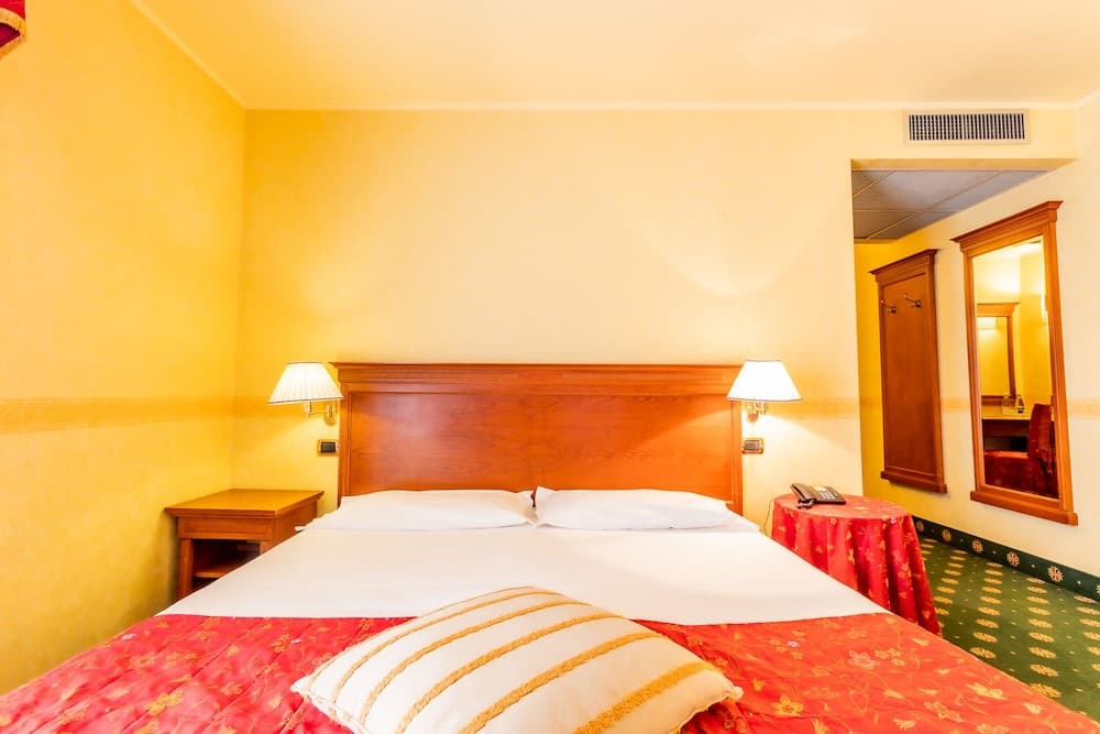 A cozy hotel room with warm yellow walls, a neatly made bed adorned with red and gold bedding, bedside lamps, and a mirror reflecting the inviting ambiance, perfect for travelers seeking comfort and relaxation.