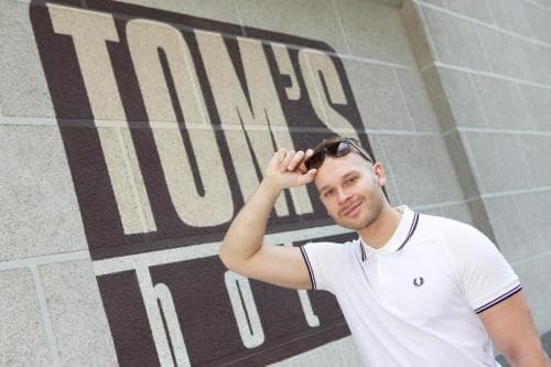 A stylish traveler poses confidently in front of a bold "TOM'S" sign on a modern brick wall, ideal for urban exploration and social media sharing.