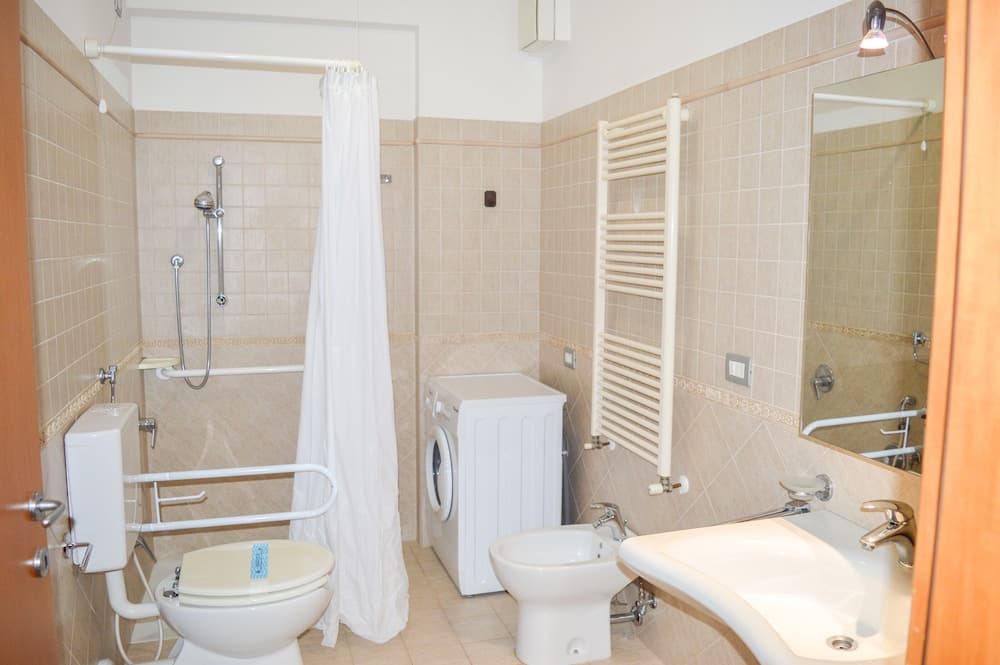 A cozy hotel bathroom features beige tiled walls, a shower with a curtain, a toilet with safety bars, a bidet, a sink with a mirror, and a towel radiator, ideal for travelers seeking comfort and accessibility.