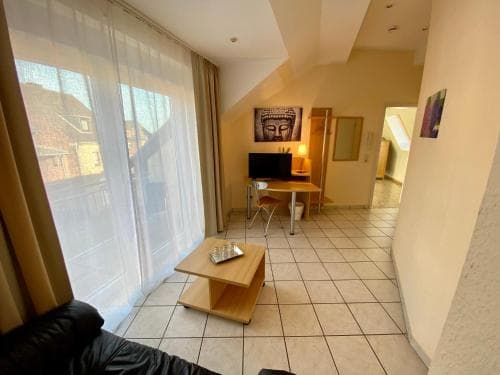 A cozy apartment interior with tiled floors, a wooden coffee table, and large windows draped in sheer curtains, exudes a tranquil ambiance ideal for travelers seeking a peaceful retreat.