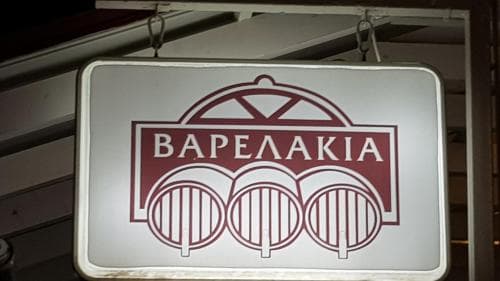 A warmly lit signboard featuring "ΒΑΡΕΛΑΚΙΑ" with wine barrel imagery sets the inviting mood of a cozy Greek taverna, perfect for travelers seeking authentic local dining experiences.
