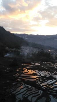 A serene sunset illuminates terraced rice fields reflecting golden hues, set against a backdrop of misty mountains and a quaint village, ideal for tranquil cultural exploration and photography.