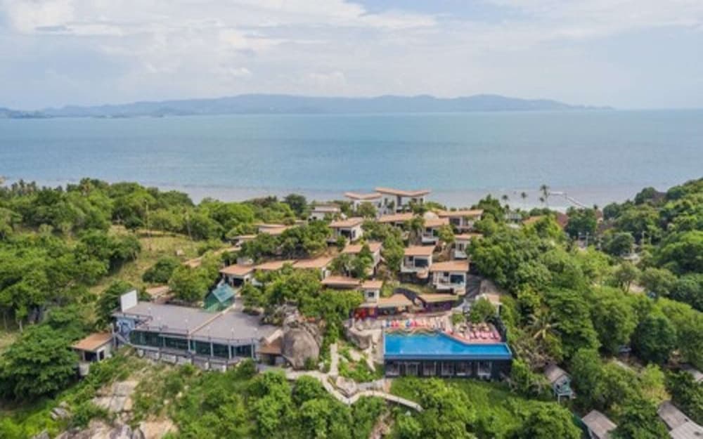 An aerial view of a luxurious hillside resort nestled among lush greenery with a vibrant pool area, overlooking the serene ocean, ideal for travelers seeking a tranquil tropical getaway.
