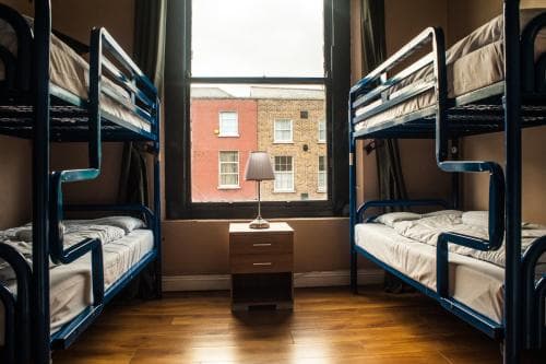 A cozy hostel room with blue bunk beds, wooden floors, and a small nightstand by the window offers travelers a budget-friendly stay in an urban setting with a warm and inviting atmosphere.