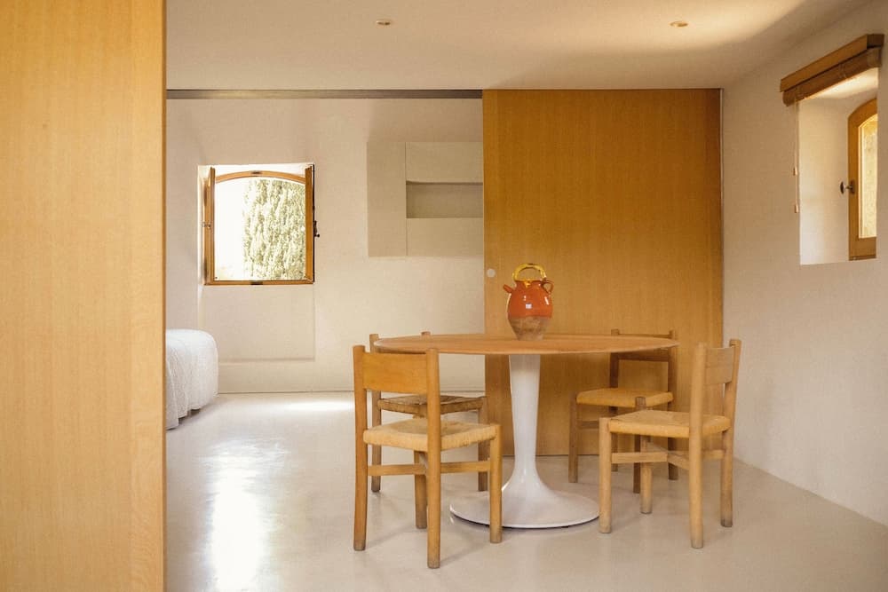 A serene minimalist bedroom with a cozy dining nook featuring wooden chairs around a round table adorned with an orange vase, ideal for travelers seeking a tranquil retreat.